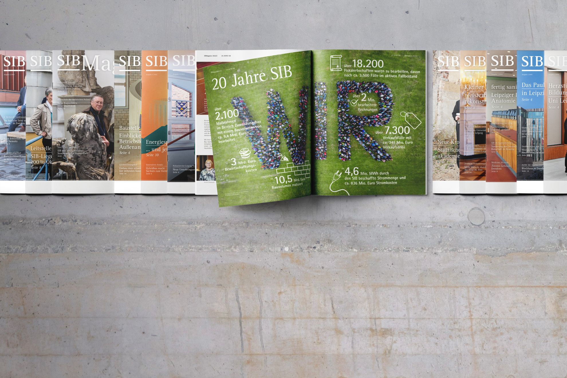 12 Ausgaben des SIB Magazines liegen in einer Reihe auf einem Rohbau-Betonhintergrund. Eines davon ist aufgeschlagen, zeigt "20 Jahre SIB" und ein "WIR" aus vielen Menschen, aus der Luft auf einer grünen Wiese fotografiert.